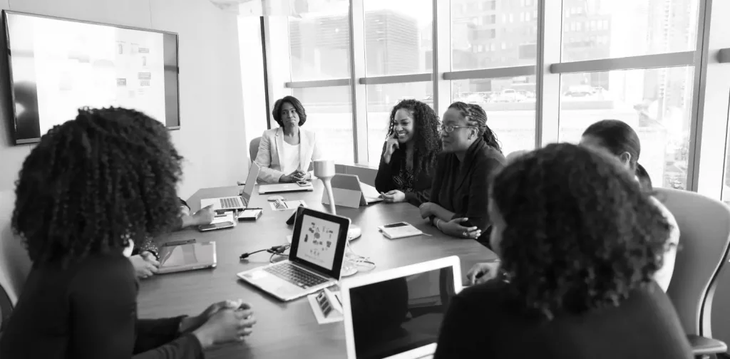 a black and white picture of women sitting together in a meeting table with laptops in front of them