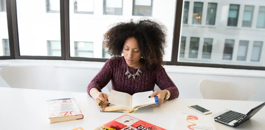 a woman with short hair sits behind a desk reading a book. the desk is cluttered with other books, laptops, and a mobile phone.