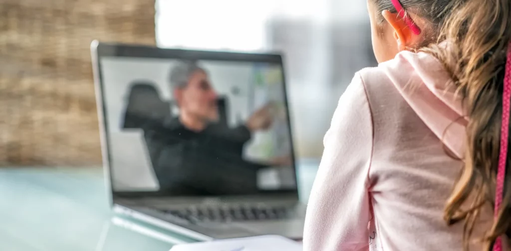 girl in pink sitting in front of macbook pro following a school lesson with male teacher speaking