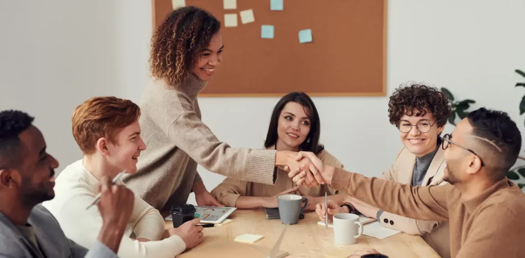 a female and a male workers are shaking hands across the table surrounded by four other colleagues.